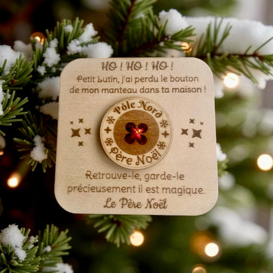 Une carte de Noël carrée en bois, suspendue à une branche de sapin de Noël saupoudrée de neige artificielle et de guirlandes lumineuses. La carte est gravée d'un message du Père Noël et comporte en son centre un bouton en bois véritable avec l'inscription "Pôle Nord" et "Père Noël" et un centre rouge brillant. La carte est entourée de petites étoiles gravées.