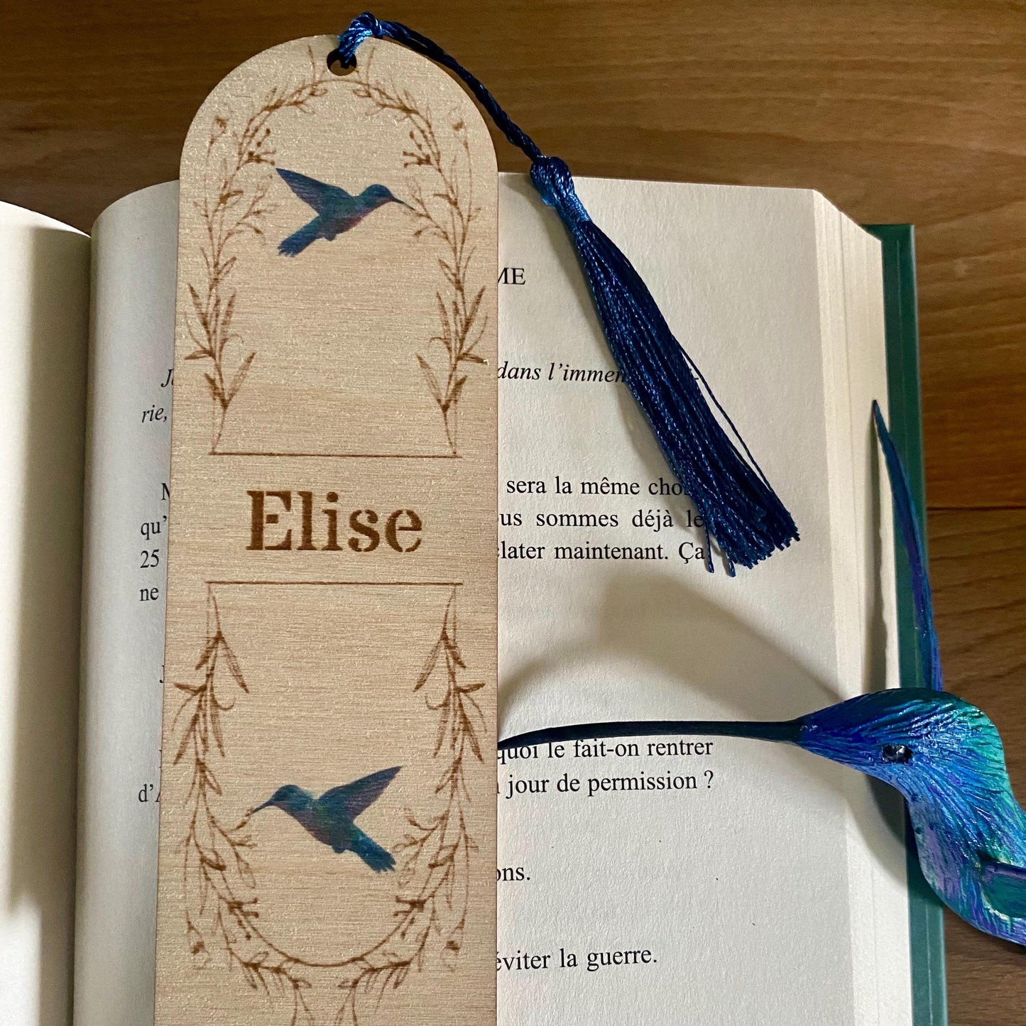 Marque-page en bois (modèle colibri) avec pompon coloré, posé à côté d'un livre ouvert.